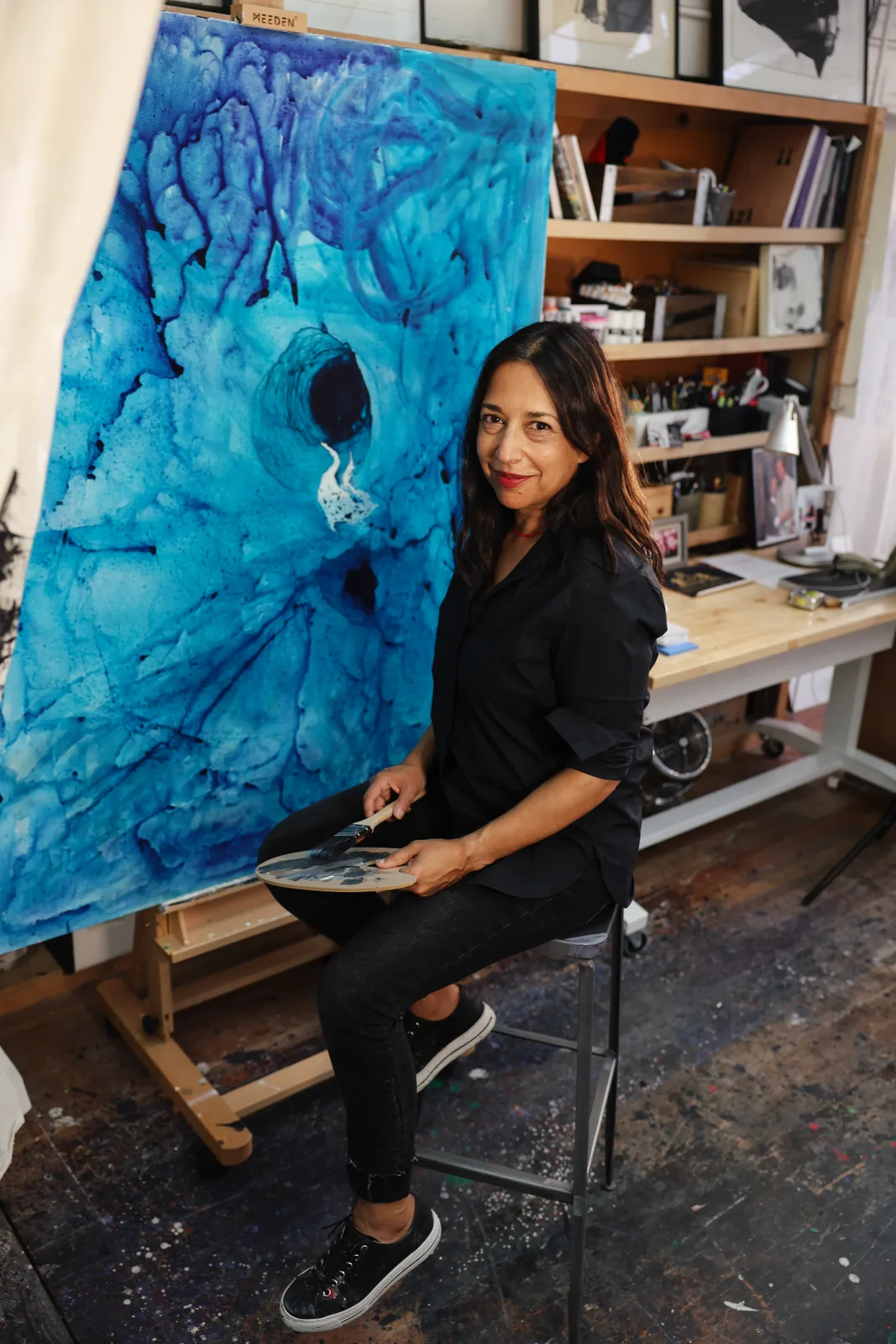 An artist seated beside her blue painting in the studio
